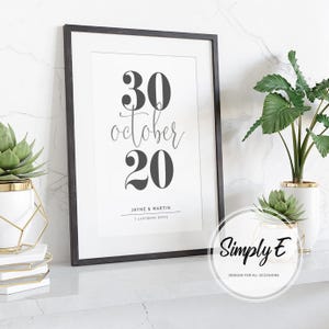 May include: A framed print with the date "30 October 20" in black against a white background. The names "Jayne & Martin" and address are below the date. The frame is black, and the print is displayed on a white surface with plants.