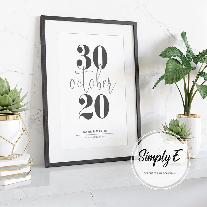 May include: A framed print with the date "30 October 20" in black against a white background. The names "Jayne & Martin" and address are below the date. The frame is black, and the print is displayed on a white surface with plants.