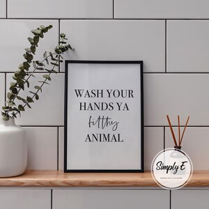 Puede incluir: Un marco negro con el texto "WASH YOUR HANDS YA filthy ANIMAL" en negro. Un jarr&oacute;n blanco con vegetaci&oacute;n y un difusor con ca&ntilde;as marrones est&aacute;n sobre una repisa de madera. El fondo es de azulejos blancos.