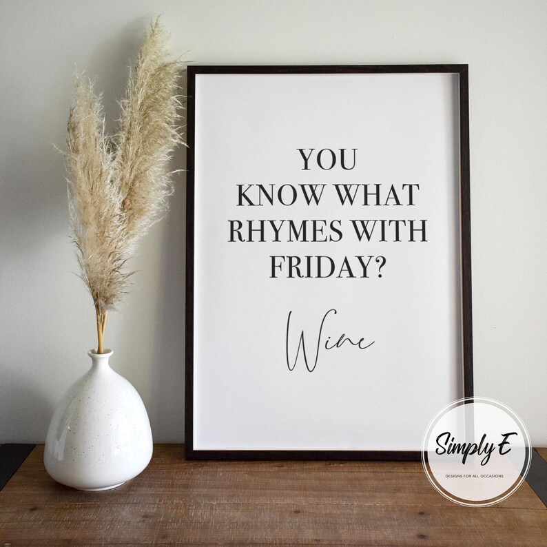 May include: A black framed print with the text "YOU KNOW WHAT RHYMES WITH FRIDAY? Wine" on a white background. A white vase with pampas grass is to the left of the frame. The frame is on a wooden surface.