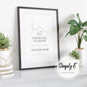 Peut inclure: Impression encadr&eacute;e avec un cadre noir, affichant l'adresse "65 Cedar Ave, Glasgow" et le texte "OUR NEW HOME" avec une date. Le design comprend une maison et des coeurs. L'impression est pr&eacute;sent&eacute;e avec des plantes dans des pots blancs et dor&eacute;s.