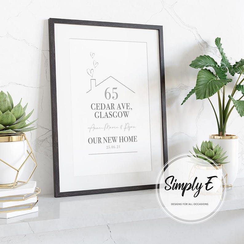 Peut inclure: Impression encadr&eacute;e avec un cadre noir, affichant l'adresse "65 Cedar Ave, Glasgow" et le texte "OUR NEW HOME" avec une date. Le design comprend une maison et des coeurs. L'impression est pr&eacute;sent&eacute;e avec des plantes dans des pots blancs et dor&eacute;s.