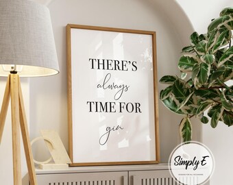 May include: A framed print with the text "THERE'S always TIME FOR gin" in black script. The print is in a light brown wooden frame and is displayed on a grey cabinet. A wooden floor lamp and a potted plant are also in the image.