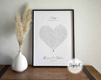 Puede incluir: Una impresión enmarcada en negro con texto blanco sobre fondo blanco. El texto es una letra de la canción "Happy" de Pharrell Williams. La impresión está personalizada con los nombres "Margaret & Stephen" y la fecha "First May 2023". La impresión está sobre una mesa de madera con un jarrón de hierba de la pampa seca.