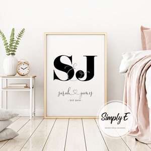 May include: A black and white printable wall art design with the initials "SJ" in a stylized font. The initials are connected by a decorative ampersand. Below the initials, the text reads "sarah & james - EST 2016 -" in a cursive font. The design is framed in a gold frame and is displayed on a white wall.