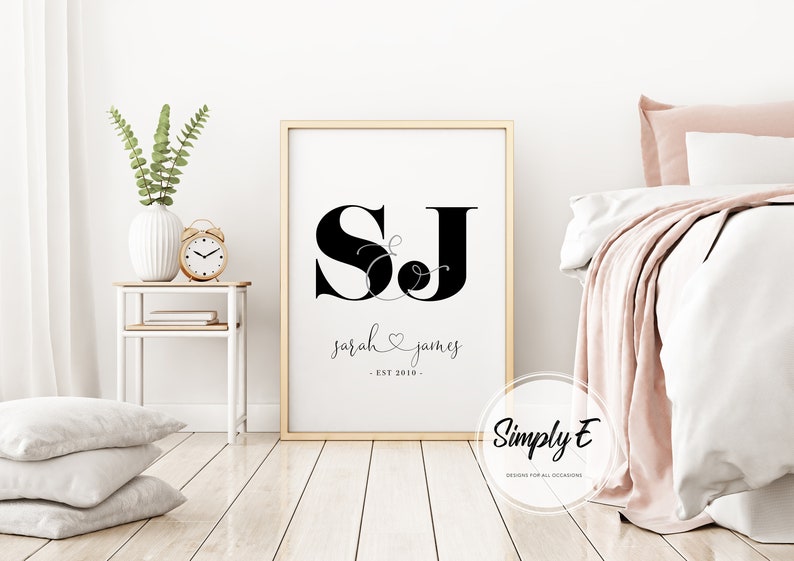 May include: A black and white printable wall art design with the initials "SJ" in a stylized font. The initials are connected by a decorative ampersand. Below the initials, the text reads "sarah & james - EST 2016 -" in a cursive font. The design is framed in a gold frame and is displayed on a white wall.