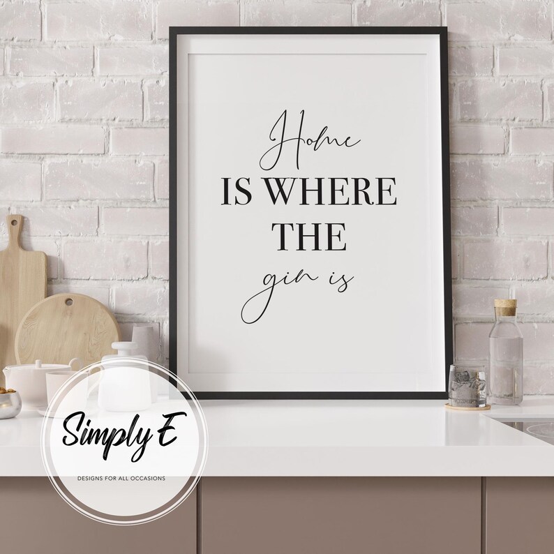 May include: A framed print with the text "Home is where the gin is" in a stylish script font. The black frame surrounds a white background. The print is displayed on a white countertop with kitchen accessories in the background.