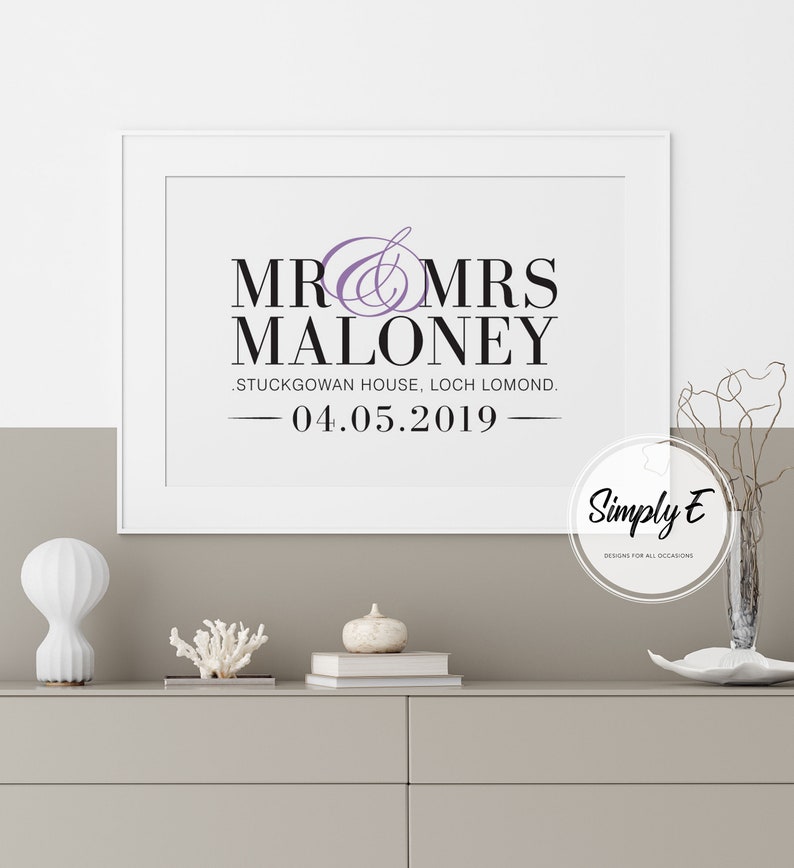 May include: A white framed print with black text that reads "Mr & Mrs Maloney, Stuckgowan House, Loch Lomond, 04.05.2019". The text is in a decorative font. The print has a purple ampersand symbol. The print is on a white background. The print is on a table with a white vase, a white pumpkin, and a white coral piece.