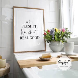 May include: A framed print with the text "ah FLUSH IT, Hush it REAL GOOD" in a bathroom setting. A vase of colorful flowers and a cup sit nearby. A wooden bath tray holds a bar of soap.