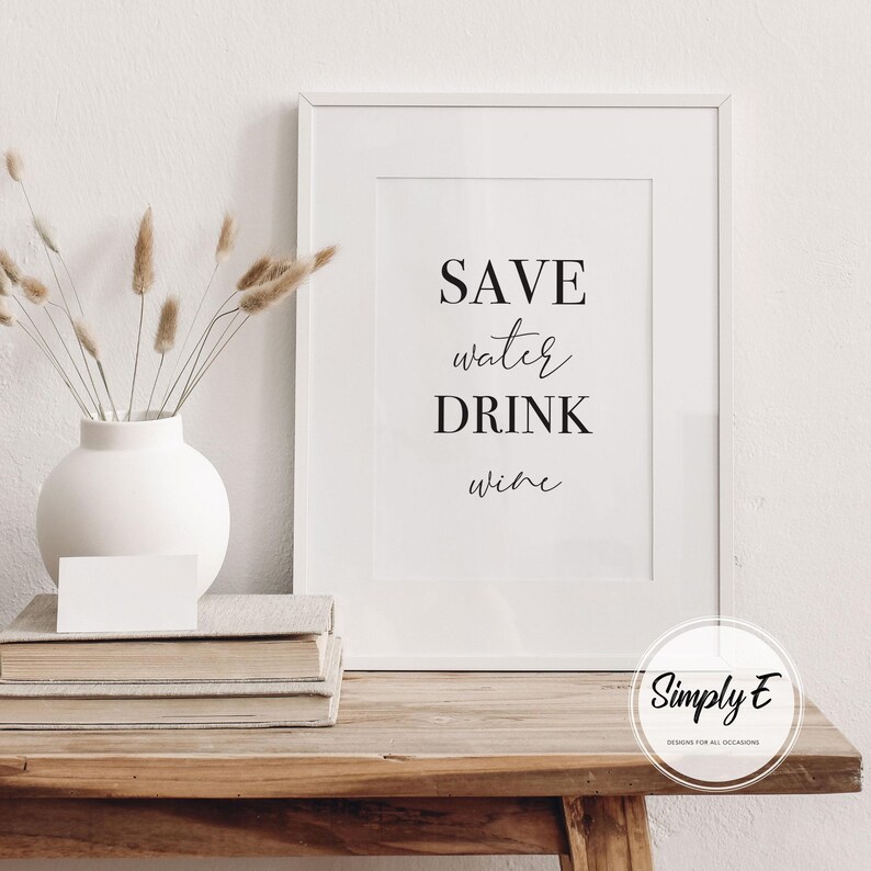 May include: A framed print with the text "SAVE water DRINK wine" in a minimalist style. The print is displayed on a wooden table, next to a white vase with dried plants and a stack of books. A small white card rests on the books.
