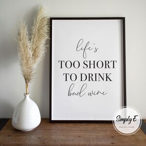 May include: A framed print with the quote "Life's too short to drink bad wine" in black script. The print is in a black frame and sits on a wooden surface. A white vase with dried pampas grass is to the left.