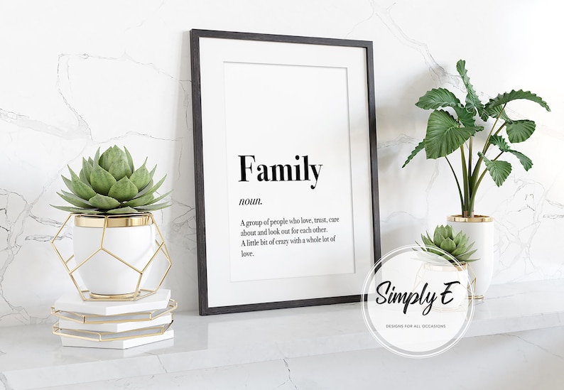 May include: A black framed print with the word "Family" in bold, followed by a definition: "A group of people who love, trust, care about and look out for each other. A little bit of crazy with a whole lot of love." The print is on a white background and is sitting on a white shelf with two potted plants.