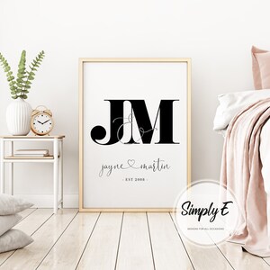 May include: A black and white printable wall art design with the initials "JM" in a large font, with a stylized ampersand and a heart symbol between the letters. The text "jayne & martin - EST 2008 -" is below the initials. The design is on a white background and is framed in a light wood frame.