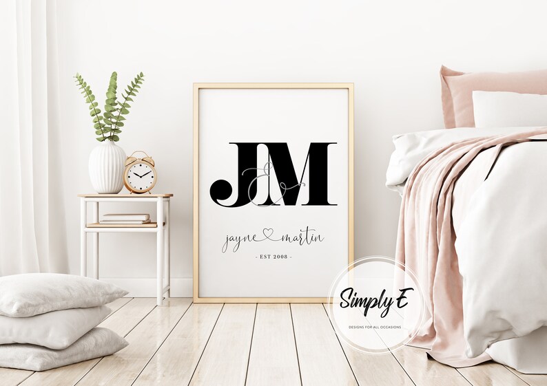 May include: A black and white printable wall art design with the initials "JM" in a large font, with a stylized ampersand and a heart symbol between the letters. The text "jayne & martin - EST 2008 -" is below the initials. The design is on a white background and is framed in a light wood frame.
