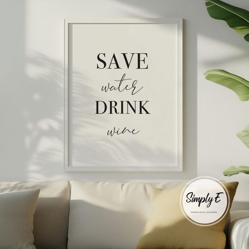 May include: A framed print with the text "SAVE water DRINK wine" in black script. The print is in a white frame and hangs on a white wall. A sofa with yellow cushions is in the foreground, with a plant in the background.