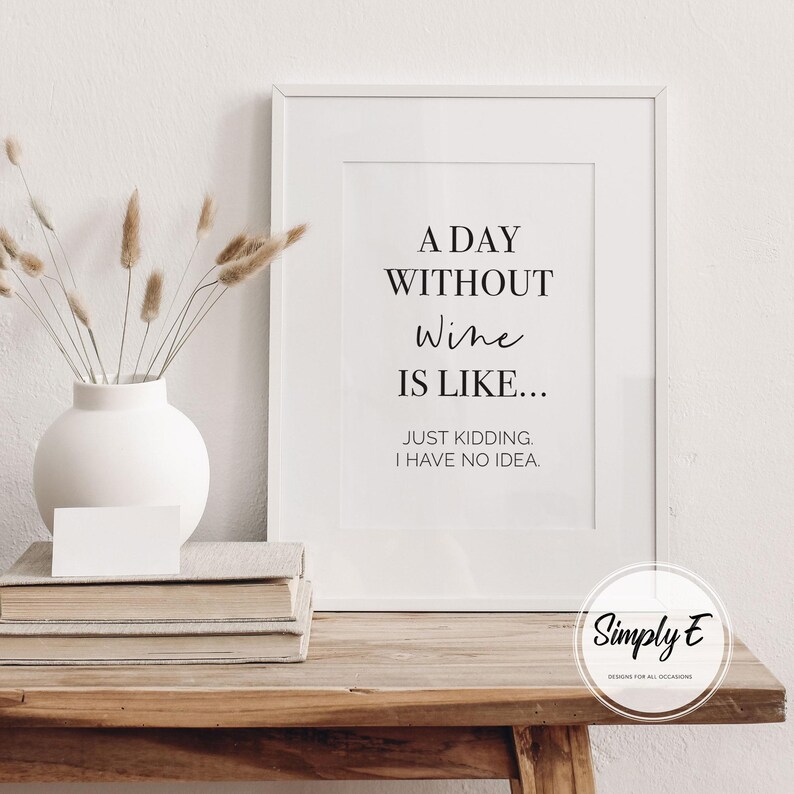 May include: A framed print with the text "A DAY WITHOUT Wine IS LIKE... JUST KIDDING. I HAVE NO IDEA." The print is displayed on a wooden table with a white vase and books. The vase contains dried flowers.
