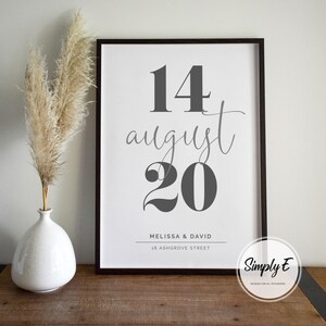 May include: A framed print with the date "14 August 20" in gray, with the names "Melissa & David" and address below. A white vase with dried pampas grass is to the left. The print is on a wooden surface.
