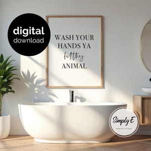May include: A rectangular wall art print in a light wood frame featuring the black text WASH YOUR HANDS YA filthy ANIMAL on a white background. The print is displayed above a white oval bathtub in a bathroom setting with a black digital download badge.