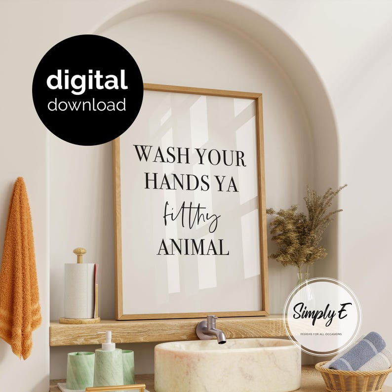 May include: A vertical rectangular wall art print in a light wood frame featuring the black text WASH YOUR HANDS YA filthy ANIMAL on a white background. The print is displayed above a bathroom sink with a faucet and a small basket of towels.