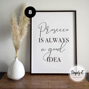 May include: A framed rectangular wall art print with a white background and black text that reads Prosecco IS ALWAYS a good IDEA. Next to the frame is a white ceramic vase containing tall, feathery beige pampas grass stems.