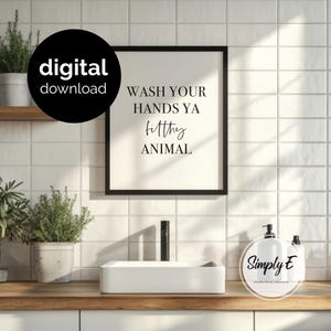 May include: A rectangular black frame containing a white print with the black text WASH YOUR HANDS YA filthy ANIMAL. The print is displayed on a white tiled wall above a white bathroom sink basin and a green potted plant in a grey pot.