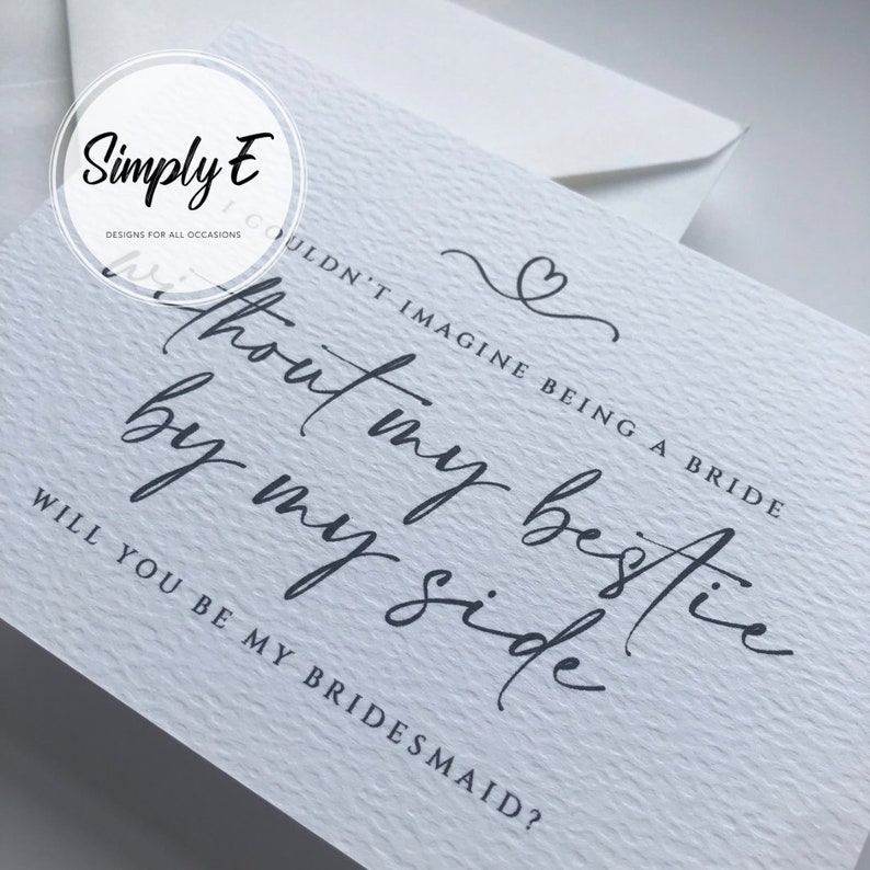 May include: A white card with black text that reads "Couldn't imagine being a bride without my bestie by my side. Will you be my bridesmaid?" The card has a simple heart design at the top.