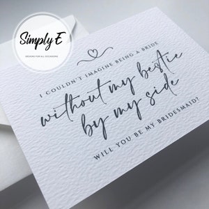 May include: A white card with a black and white design. The card says "I couldn't imagine being a bride without my bestie by my side. Will you be my bridesmaid?"
