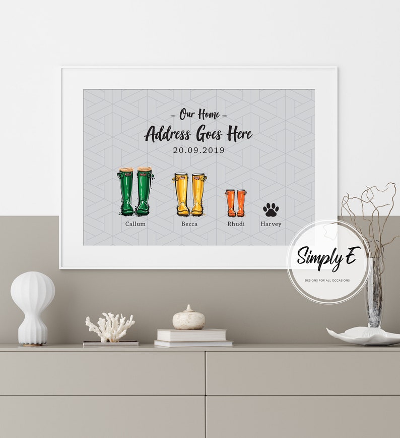 May include: A framed print with a gray background and a geometric pattern. The print features a family of four, represented by colorful rain boots, with the names Callum, Becca, Rhudi, and Harvey. The text "Our Home - Address Goes Here 20.09.2019" is at the top of the print. The print is framed in white and has a white mat.