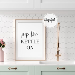 Peut inclure: Une affiche de cuisine encadr&eacute;e avec un fond blanc et un texte noir qui dit "pop the kettle on". L'affiche est accroch&eacute;e &agrave; un mur blanc avec un motif de carreaux hexagonaux blancs. Un vase rose avec des fleurs roses est sur un comptoir blanc.