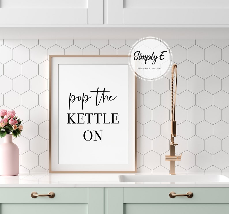 Peut inclure: Une affiche de cuisine encadr&eacute;e avec un fond blanc et un texte noir qui dit "pop the kettle on". L'affiche est accroch&eacute;e &agrave; un mur blanc avec un motif de carreaux hexagonaux blancs. Un vase rose avec des fleurs roses est sur un comptoir blanc.