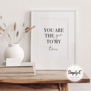 Puede incluir: Impresi&oacute;n enmarcada en blanco con el texto "YOU ARE THE gin TO MY tonic" en negro. Sobre una mesa de madera, con un jarr&oacute;n blanco con plantas secas y libros. Una tarjeta blanca descansa sobre los libros.