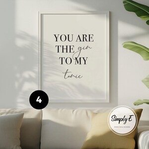 May include: A rectangular wall art print in a white frame featuring the black text YOU ARE THE gin TO MY tonic on a cream background. The print is displayed above a sofa with a yellow cushion and a black circular icon with the number 4.