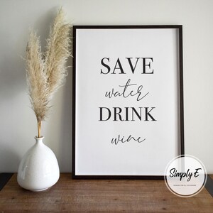 May include: A framed print with the words "SAVE water DRINK wine" in black script. A white vase with pampas grass is to the left of the frame. The frame is on a wooden surface.