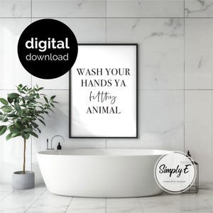 May include: A rectangular black-framed wall art print featuring the black text WASH YOUR HANDS YA filthy ANIMAL on a white background. The print is displayed above a white bathtub. A black circular badge with the white text digital download is visible.