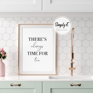 Op de afbeelding: Een ingelijst print met de tekst "THERE'S always TIME FOR tea" in een wit frame op een wit aanrecht met een roze vaas met bloemen en een gouden kraan.