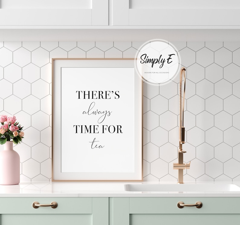 Op de afbeelding: Een ingelijst print met de tekst "THERE'S always TIME FOR tea" in een wit frame op een wit aanrecht met een roze vaas met bloemen en een gouden kraan.