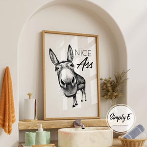 May include: A framed black and white print featuring a donkey with the text "NICE Ass". The artwork is displayed in a bathroom setting, with a wooden frame and a neutral color palette.