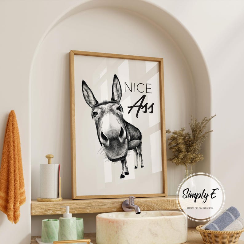 May include: A framed black and white print featuring a donkey with the text "NICE Ass". The artwork is displayed in a bathroom setting, with a wooden frame and a neutral color palette.