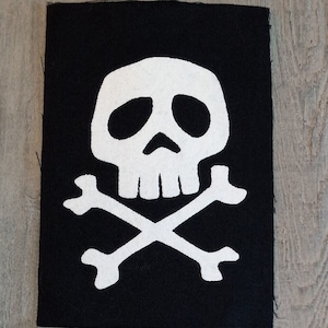 May include: Black rectangular patch featuring a white skull and crossbones design. The skull has large eye sockets and a prominent nose. The crossbones are positioned beneath the skull. The patch is likely for clothing or accessories.