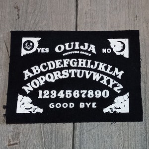 May include: Black Ouija board patch with white lettering and symbols. The patch includes the alphabet, numbers, and the words "Yes," "No," and "Good Bye." The words "Ouija Mystifying Oracle" are also present.