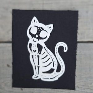 May include: A white cat skeleton design on a black square. The cat is sitting with its tail curled. The design is a cut-out, showing the cat's skeletal structure. The background is a light grey wooden surface.