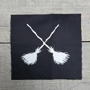 May include: A black square patch featuring a white graphic of two crossed witch brooms. The brooms are depicted with detailed bristles and crossed at the handles. The patch is set against a weathered wooden surface.