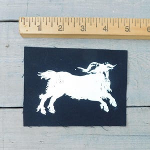 Op de afbeelding: Een zwarte stoffen patch met een witte illustratie van een geit in een rennende pose. De patch is rechthoekig en is ongeveer 7.6 cm breed. Een houten liniaal is boven de patch geplaatst.