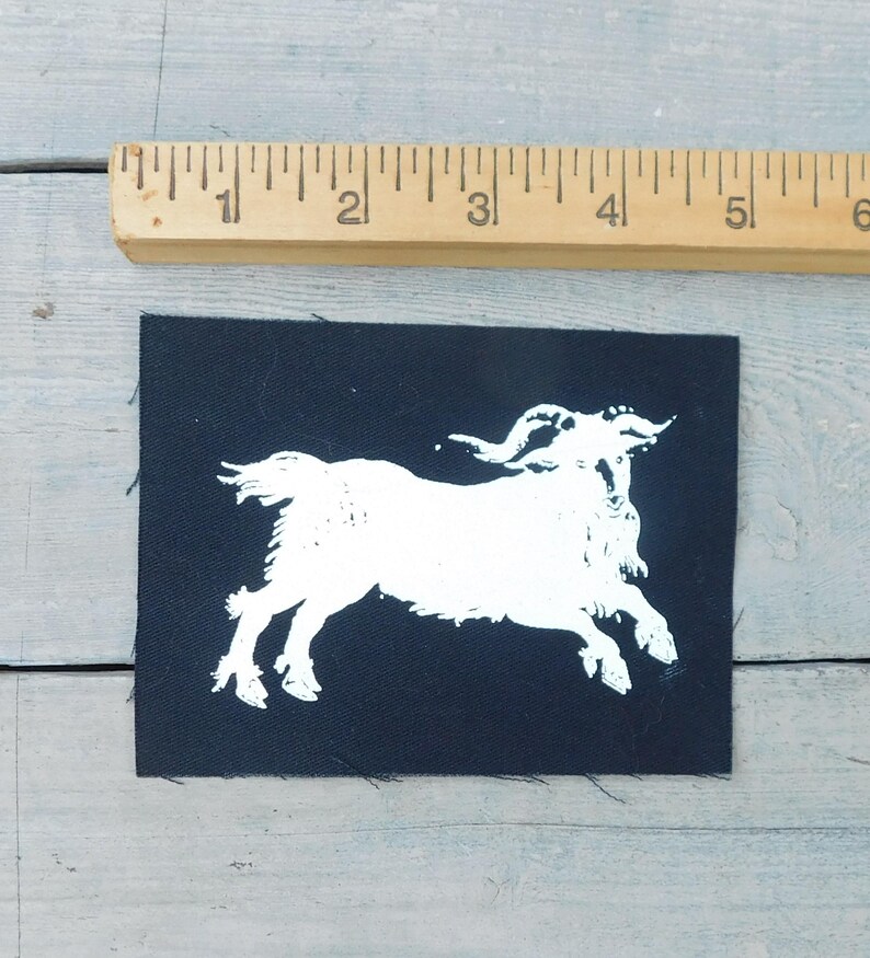 Op de afbeelding: Een zwarte stoffen patch met een witte illustratie van een geit in een rennende pose. De patch is rechthoekig en is ongeveer 7.6 cm breed. Een houten liniaal is boven de patch geplaatst.