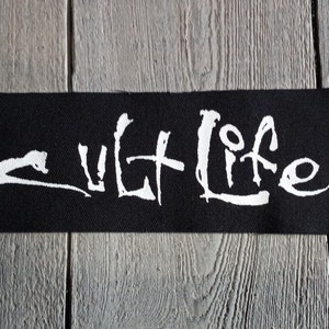 May include: A black rectangular patch featuring the white text "cult LIFE". The patch is made of woven fabric and is displayed on a grey wooden surface.