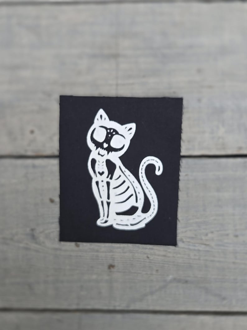 May include: A white cat skeleton design on a black square. The cat is sitting with its tail curled. The design is a cut-out, showing the cat's skeletal structure. The background is a light grey wooden surface.