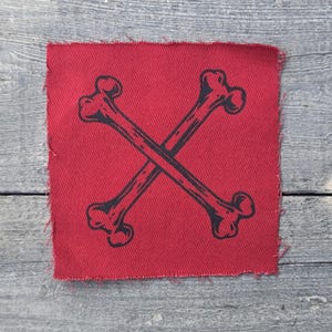 May include: A square fabric patch in a deep red color featuring a black screen-printed graphic of two crossed bones. The patch has raw, frayed edges along all four sides, giving it a textured appearance against the background surface.