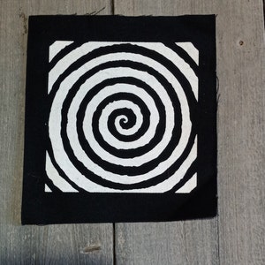 May include: A black square patch with a white spiral pattern. The spiral is contained within a white square, surrounded by a black border. The patch appears to be fabric and is placed on a wooden surface.