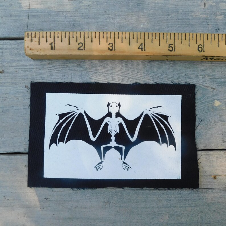 Op de afbeelding: Een zwart-wit patch met een vleermuisskeletontwerp. De vleermuis heeft uitgespreide vleugels op een witte achtergrond, binnen een rechthoekige zwarte rand. Een liniaal is boven de patch geplaatst. De patch is ongeveer 15 cm lang.