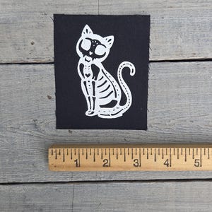 May include: A black square patch with a white cat skeleton design. The cat is sitting upright with its tail curled. The patch is on a grey wooden surface next to a wooden ruler.